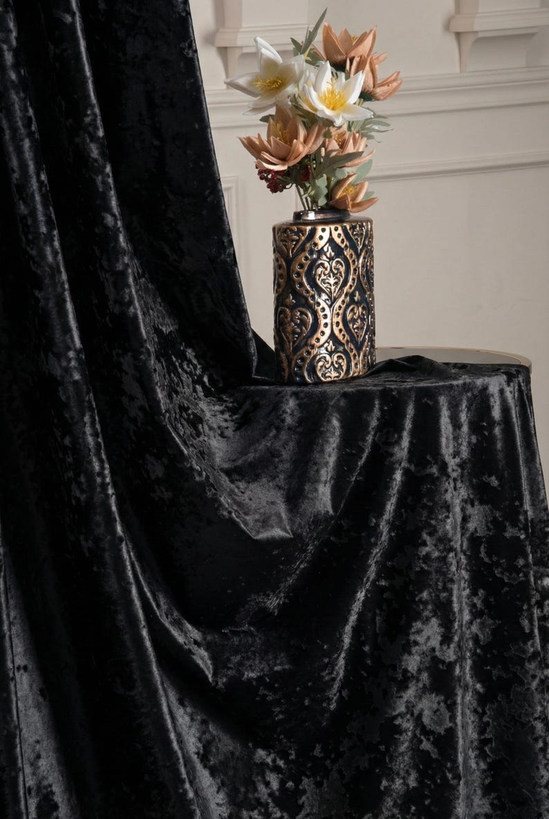 K&ouml;nnte beinhalten: Ein schwarzer Vorhang aus Knitter-Samt mit tiefem, strukturiertem Flor. Neben dem Stoff steht eine zylindrische Vase mit einem metallisch goldenen und schwarzen Ornamentmuster, die einen Strau&szlig; aus wei&szlig;en und pfirsichfarbenen Kunstblumen enth&auml;lt.