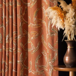 Puede incluir: Cortina de color terracota con un patrón abstracto repetido en blanco, dorado y verde oliva. La cortina está drapeada, con pliegues verticales. Un jarrón de bronce con hierba ornamental seca se encuentra sobre una pequeña mesa de madera.