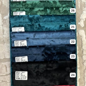 K&ouml;nnte beinhalten: Eine Stoffmusterkarte mit sieben horizontalen Streifen aus Knitter-Samt in den Farben Hellblau, Gr&uuml;n, T&uuml;rkis, Blau, Dunkelblau, Schwarz und Rot. Jeder Streifen ist mit einem wei&szlig;en Etikett versehen, das DESIGN:TIBET, WIDTH: 300 cm und die Nummern 27, 26, 21, 22, 23, 24, 29 zeigt.