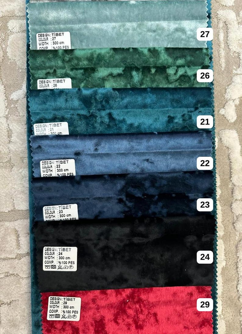 K&ouml;nnte beinhalten: Eine Stoffmusterkarte mit sieben horizontalen Streifen aus Knitter-Samt in den Farben Hellblau, Gr&uuml;n, T&uuml;rkis, Blau, Dunkelblau, Schwarz und Rot. Jeder Streifen ist mit einem wei&szlig;en Etikett versehen, das DESIGN:TIBET, WIDTH: 300 cm und die Nummern 27, 26, 21, 22, 23, 24, 29 zeigt.