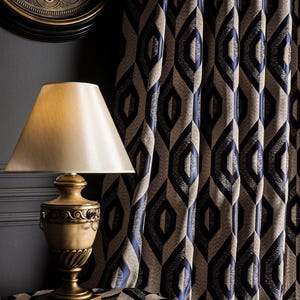 May include: A gold-toned table lamp with a cream-colored lampshade sits on a table covered with a geometric patterned fabric. Behind it, navy blue and tan curtains with a repeating diamond design hang. A clock is visible in the background.