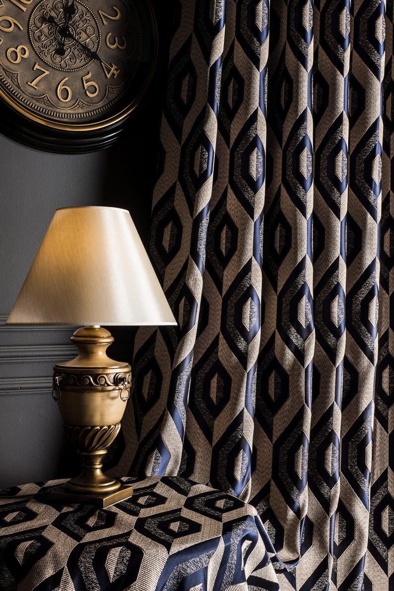 May include: A gold-toned table lamp with a cream-colored lampshade sits on a table covered with a geometric patterned fabric. Behind it, navy blue and tan curtains with a repeating diamond design hang. A clock is visible in the background.