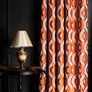 May include: Orange and cream geometric patterned curtains. The curtains are hanging next to a black side table with a lamp. The lamp has a beige lampshade and a decorative base. The curtains have a repeating diamond pattern.