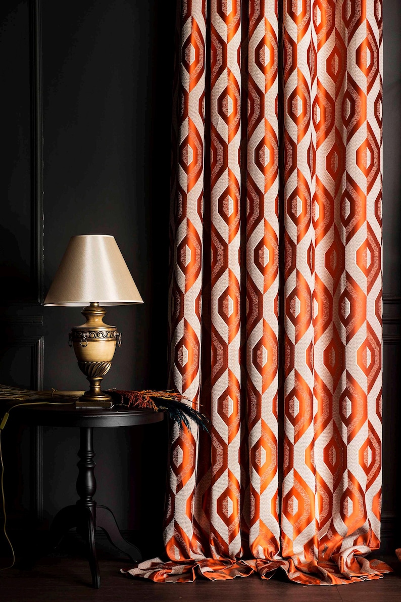 May include: Orange and cream geometric patterned curtains. The curtains are hanging next to a black side table with a lamp. The lamp has a beige lampshade and a decorative base. The curtains have a repeating diamond pattern.