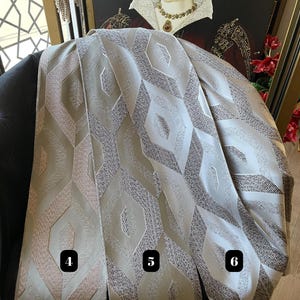 May include: Three fabric swatches with geometric patterns in shades of beige, gray, and brown. The swatches are draped over a black chair, with a framed portrait in the background. The fabric has a textured appearance, with a repeating diamond-like design.