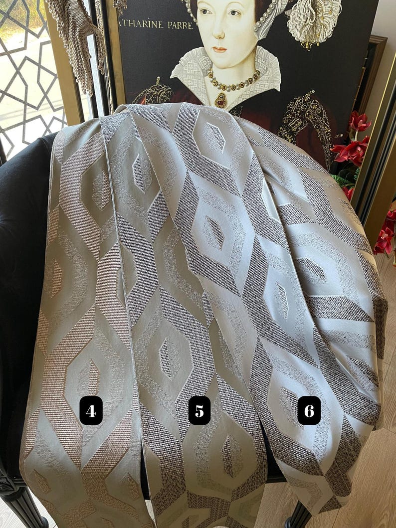 May include: Three fabric swatches with geometric patterns in shades of beige, gray, and brown. The swatches are draped over a black chair, with a framed portrait in the background. The fabric has a textured appearance, with a repeating diamond-like design.