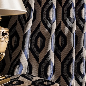 May include: A decorative gold-toned lamp with a cream-colored lampshade sits on a patterned table. Behind the lamp, textured curtains with a geometric diamond pattern in navy blue and tan create a sophisticated backdrop.