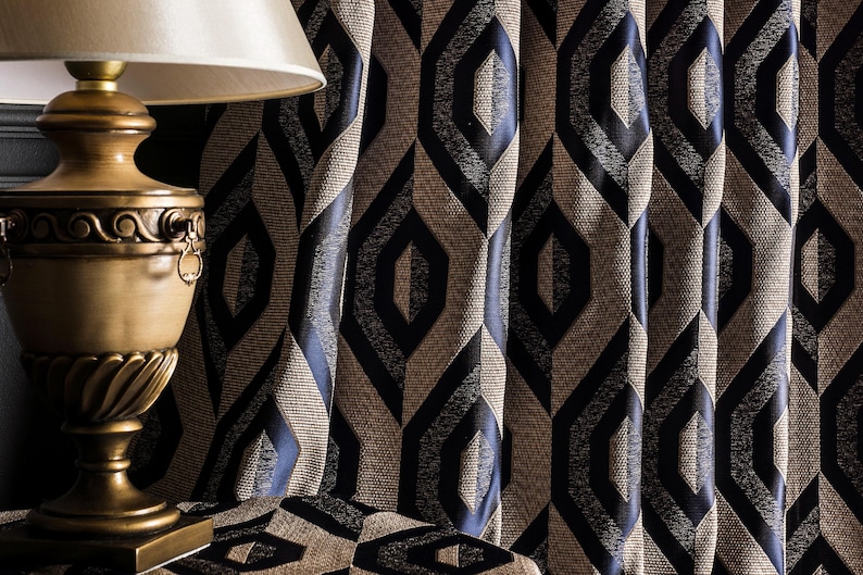 May include: A decorative gold-toned lamp with a cream-colored lampshade sits on a patterned table. Behind the lamp, textured curtains with a geometric diamond pattern in navy blue and tan create a sophisticated backdrop.