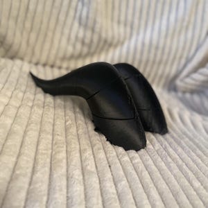 Op de afbeelding: Een paar zwarte, gebogen hoorns gemaakt van glad, hard materiaal. De hoorns liggen op een wit, pluche oppervlak.