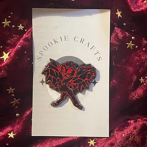 May include: A red enamel pin featuring a black and red cartoon creature with wings. The pin says "SPOOKJE CRAFTS" in a circle around the creature.