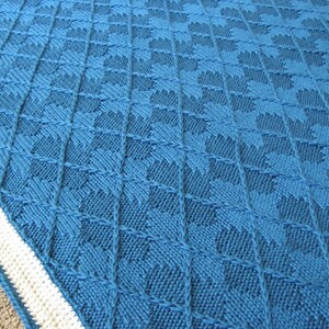 Op de afbeelding: Een blauwe gebreide deken met een witte rand. De deken heeft een herhalend patroon van kleine, ruitvormige ontwerpen.