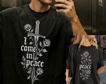 Può includere: Una t-shirt nera a maniche corte con una stampa grafica bianca raffigurante una spada intrecciata con rose. Il testo "I come in peace" è stampato in un carattere in stile gotico accanto alla spada, centrato sulla parte anteriore del capo.