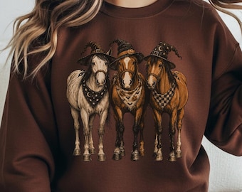 Può includere: Una felpa girocollo color marrone cioccolato con una stampa grafica di tre cavalli in piedi uno accanto all'altro. Ogni cavallo indossa un cappello da strega a punta e una bandana fantasia al collo. I cavalli sono in tonalità bianco, beige e marrone.
