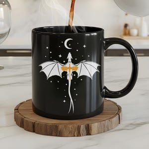 Op de afbeelding: Een zwarte keramische mok met een drakenontwerp. De draak heeft witte vleugels en een gouden lichaam, met een maan en sterren erboven. Koffie wordt in de mok gegoten, die op een houten onderzetter staat.