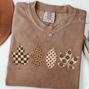 May include: A brown short-sleeve t-shirt featuring four printed leaves across the front. The leaves display distinct patterns: a black and tan checkerboard, a brown leopard print, black polka dots, and black flowers. The inner label reads Comfort Colors.