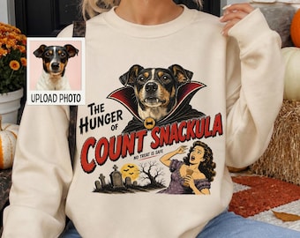 Può includere: Una felpa girocollo color sabbia con una stampa grafica di un cane vestito da vampiro, il testo The Hunger of Count Snackula No Treat Is Safe e un'illustrazione di una persona e una scena di un cimitero.