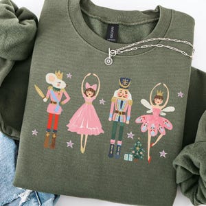 Könnte beinhalten: Ein olivgrünes Sweatshirt mit einem Nussknacker-Themen-Design. Der Aufdruck zeigt einen Mäusekönig, eine Ballerina, einen Nussknackersoldaten und eine Fee, zusammen mit einem kleinen Weihnachtsbaum und Sternen. Eine silberne Halskette ist über dem Sweatshirt drapiert.