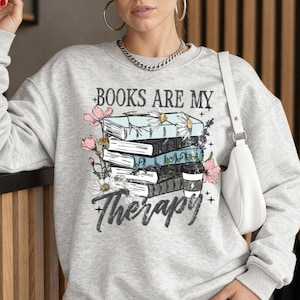 Op de afbeelding: Een lichtgrijze sweatshirt met de tekst "BOOKS ARE MY Therapy". De afbeelding toont een stapel boeken met roze en witte bloemen. De sweatshirt is een casual stijl.
