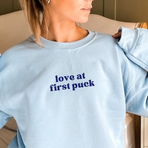 Op de afbeelding: Lichtblauwe trui met de tekst "love at first puck" in donkerblauw.