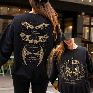 May include: Black sweatshirt with gold-colored bat wing designs and text. The back features three bat wing illustrations with names: Aerial, Cassian, and Rhysand. The front displays "THE BAT BOYS" with bat wings and names: Rhysand, Azriel, and Cassian.