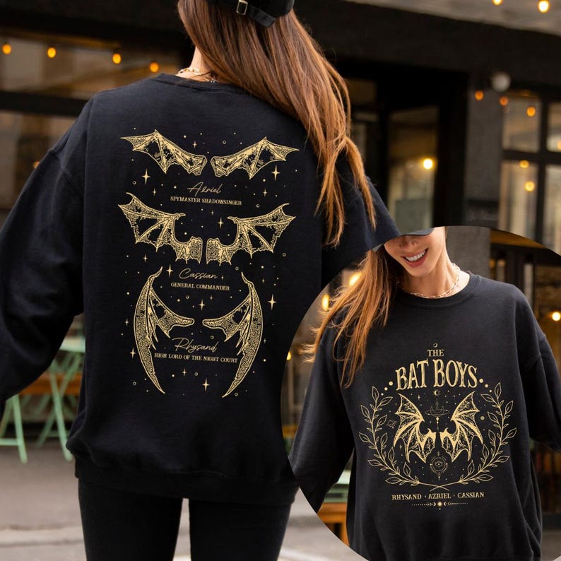 May include: Black sweatshirt with gold-colored bat wing designs and text. The back features three bat wing illustrations with names: Aerial, Cassian, and Rhysand. The front displays "THE BAT BOYS" with bat wings and names: Rhysand, Azriel, and Cassian.