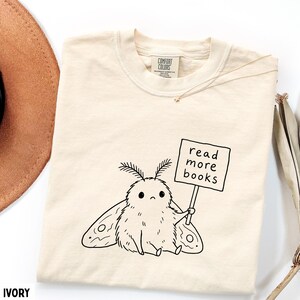 May include: An ivory t-shirt featuring a black line art illustration of a fluffy moth with antennae and large wings. The moth holds a rectangular sign that reads "read more books" in lowercase letters. The shirt has a Comfort Colors brand label.