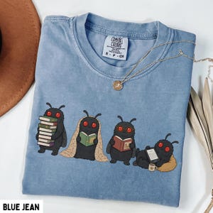 Puede incluir: Una camiseta color azul vaquero con un gr&aacute;fico de cuatro criaturas negras parecidas a polillas con ojos rojos leyendo libros. Una polilla sostiene una pila de libros, otra est&aacute; envuelta en una manta, otra lleva gafas y otra descansa sobre un coj&iacute;n.
