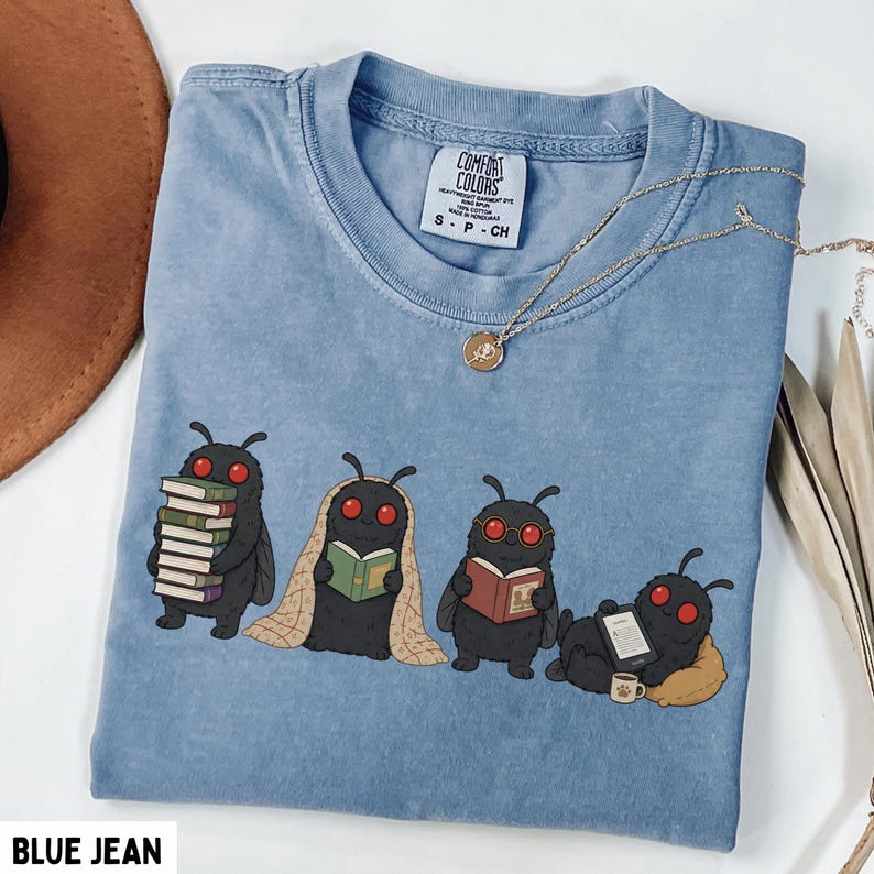 Puede incluir: Una camiseta color azul vaquero con un gr&aacute;fico de cuatro criaturas negras parecidas a polillas con ojos rojos leyendo libros. Una polilla sostiene una pila de libros, otra est&aacute; envuelta en una manta, otra lleva gafas y otra descansa sobre un coj&iacute;n.