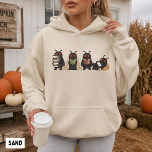 Puede incluir: Una sudadera con capucha de color arena que presenta un estampado gr&aacute;fico de cuatro criaturas negras similares a polillas con ojos rojos. Las criaturas aparecen realizando diversas actividades: sosteniendo libros, leyendo o descansando.