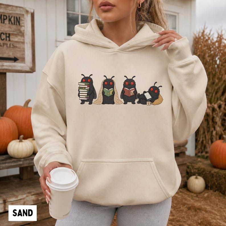 Puede incluir: Una sudadera con capucha de color arena que presenta un estampado gr&aacute;fico de cuatro criaturas negras similares a polillas con ojos rojos. Las criaturas aparecen realizando diversas actividades: sosteniendo libros, leyendo o descansando.