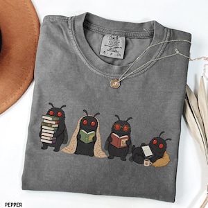 Puede incluir: Una camiseta gris carb&oacute;n con un gr&aacute;fico de cuatro criaturas negras con ojos rojos leyendo libros y usando un lector electr&oacute;nico. La camiseta tiene una etiqueta Comfort Colors en el cuello, talla S, y se acompa&ntilde;a de un collar de tono plateado.