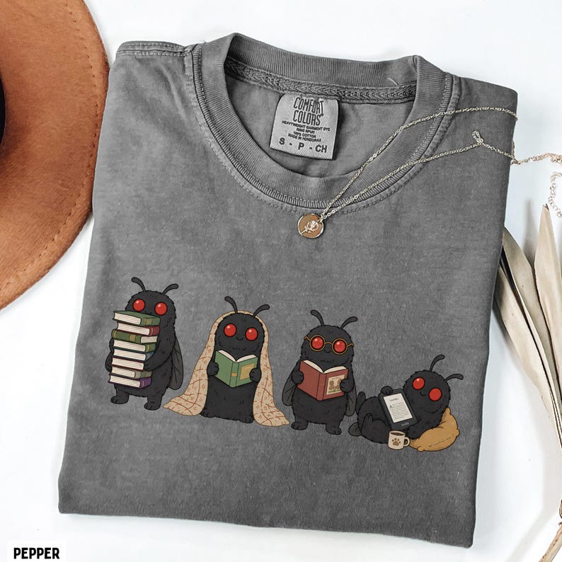 Puede incluir: Una camiseta gris carb&oacute;n con un gr&aacute;fico de cuatro criaturas negras con ojos rojos leyendo libros y usando un lector electr&oacute;nico. La camiseta tiene una etiqueta Comfort Colors en el cuello, talla S, y se acompa&ntilde;a de un collar de tono plateado.