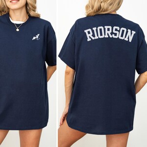 May include: Navy blue t-shirt with a small white graphic on the front and the word "RIORSON" printed in white on the back. The shirt has short sleeves and a crew neck. The model is wearing a white undershirt.