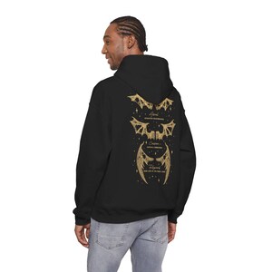 May include: Black hoodie with a gold graphic of three sets of stylized wings and text. The text includes the words "Astral", "Celestial Formations", and "Regina". The hoodie has a hood and a relaxed fit.
