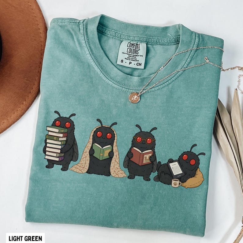 Puede incluir: Una camiseta verde claro con un gr&aacute;fico de cuatro criaturas negras parecidas a polillas con ojos rojos leyendo libros. Una polilla lleva una pila de libros, otra lee bajo una manta, otra lleva gafas y otra lee en una tableta.