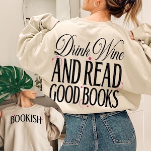 Könnte beinhalten: Beiges Sweatshirt mit dem Text "Drink Wine AND READ GOOD BOOKS" in schwarzer Schrift und Blockbuchstaben. Das Sweatshirt hat auch rosa Weinflecken-Grafiken. Ein weiteres Sweatshirt im Bild trägt die Aufschrift "BOOKISH" in schwarzen Buchstaben.