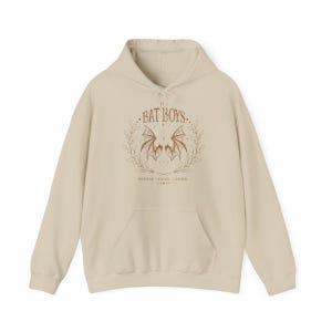 May include: A cream-colored hoodie with a front pocket and drawstring hood. The front features a brown graphic with the text "THE BAT BOYS" above a bat wing design, surrounded by a wreath. Below the design is additional text.