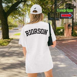 May include: A white t-shirt with the name "RIORSON" in black block letters on the back. A white baseball cap with a black dog silhouette is worn. A black shoulder bag is also visible.