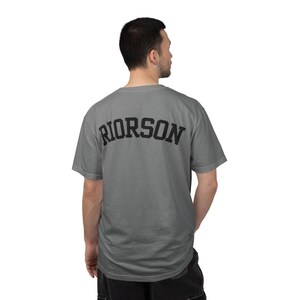 May include: A grey t-shirt with the word "RIORSON" printed in black across the upper back. The shirt has short sleeves and a crew neck. The person wearing the shirt is facing away from the camera.