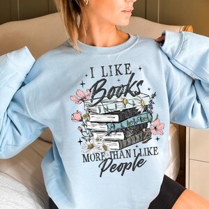 Op de afbeelding: Lichtblauw sweatshirt met de tekst "I Like Books More Than I Like People". De afbeelding toont een stapel boeken met roze en witte bloemaccenten. Het sweatshirt is effen lichtblauw van kleur.