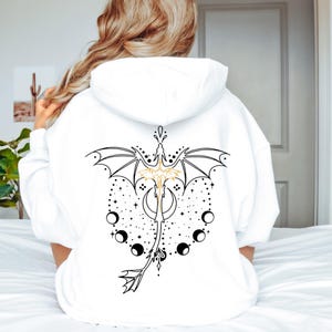 Può includere: Felpa con cappuccio bianca con un disegno di drago nero e oro sul retro. Il drago è raffigurato con ali, una luna crescente e fasi lunari. La felpa ha un cappuccio e maniche lunghe.