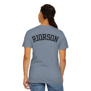 May include: A pale blue t-shirt featuring the word "RIORSON" in black, block lettering on the back. The shirt has a crew neck and is likely made from a soft fabric.