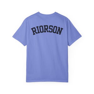 May include: A pale blue t-shirt with the word "RIORSON" printed in black, arched block letters across the back. The shirt has short sleeves and a crew neck. The fabric appears soft.