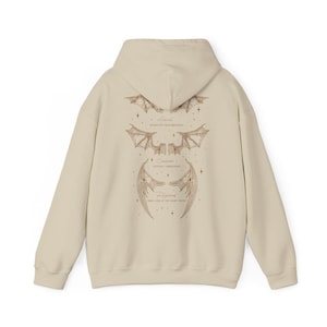 May include: A beige hoodie with a detailed illustration of three sets of stylized wings in shades of brown. The wings are labeled "Astral", "Cassian", and "Feyre". The hoodie has a drawstring hood and a kangaroo pocket.