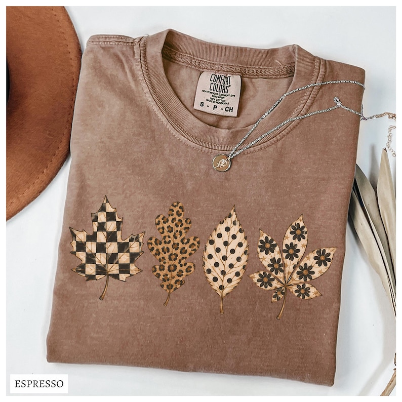 May include: A brown short-sleeve t-shirt featuring four printed leaves across the front. The leaves display distinct patterns: a black and tan checkerboard, a brown leopard print, black polka dots, and black flowers. The inner label reads Comfort Colors.