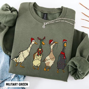 Könnte beinhalten: Ein militärgrüner Rundhals-Pullover mit einem Grafikdruck von vier Enten, die mit Weihnachtsbeleuchtung geschmückt sind. Die Enten tragen festliche Accessoires wie Weihnachtsmützen und einen rot-grünen Schal. Das Sweatshirt hat ein Gildan-Etikett.