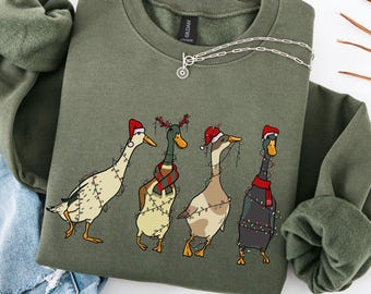 Puede incluir: Una sudadera de cuello redondo color verde militar con un estampado gráfico de cuatro patos decorados con luces navideñas. Los patos llevan accesorios festivos, incluyendo gorros de Papá Noel y una bufanda roja y verde. Tiene etiqueta Gildan.