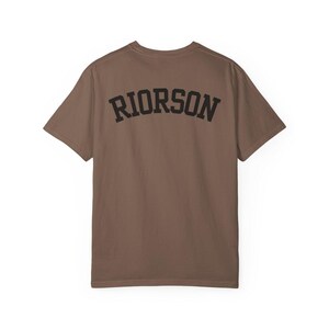 May include: A brown t-shirt featuring the word "RIORSON" in black, arched lettering across the back. The shirt has a crew neck and short sleeves. The material looks like a soft cotton blend.