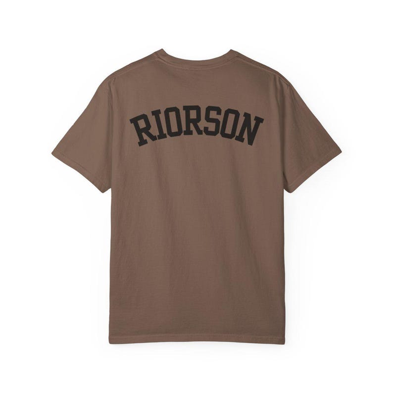 May include: A brown t-shirt featuring the word "RIORSON" in black, arched lettering across the back. The shirt has a crew neck and short sleeves. The material looks like a soft cotton blend.
