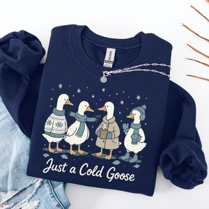 Könnte beinhalten: Marineblaues Sweatshirt mit einer Grafik von vier Comic-Gänsen, die winterlich gekleidet sind. Die Gänse tragen Pullover, Schals, Mäntel und Mützen. Der Text "Just a Cold Goose" ist darunter aufgedruckt. Eine silberne Halskette liegt auf dem Sweatshirt.